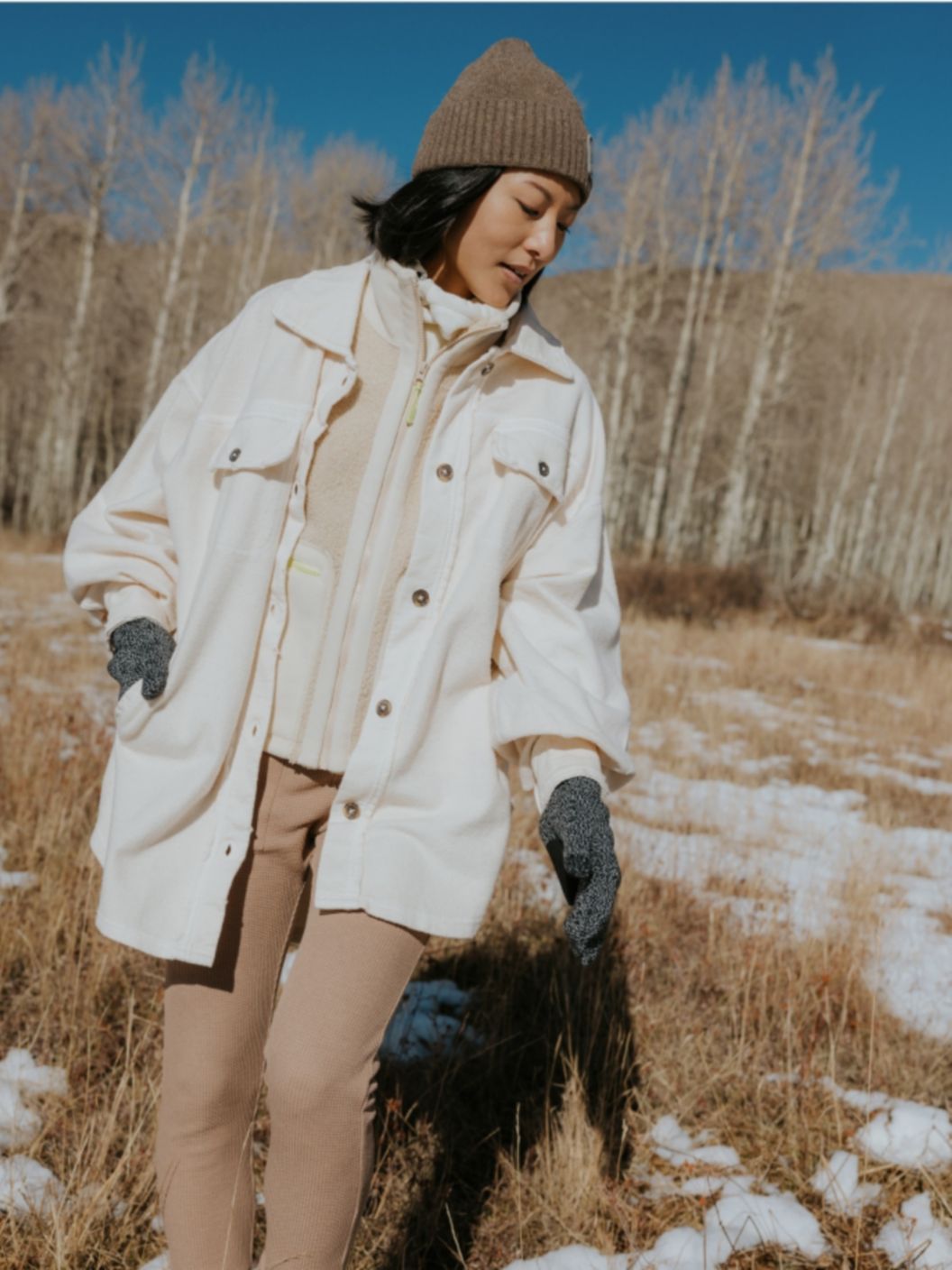   A woman walking in a snowy field wears a monochrome layered outfit, including leggings, fleece sweater, felt coat, and knit hat. 